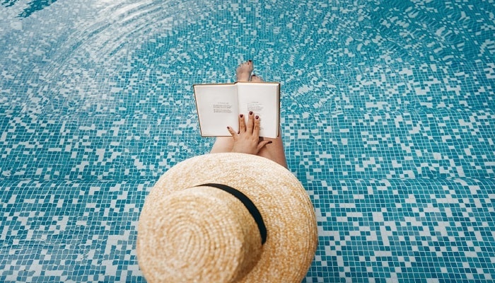 A person wearing a straw hat sitting in a pool reading a book.