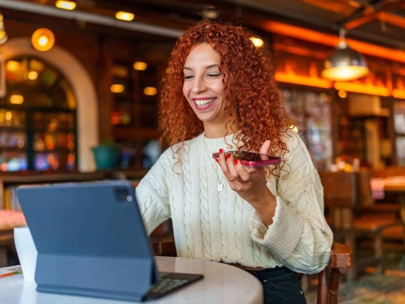 Frau mit roten und gelockten haaren redet am Telefon während sie vor einem Laptop im Cafe sitzt.