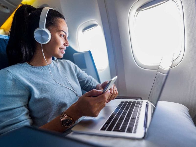 Frau sitz im Flugzeug während sie mit Kopfhörern Musik hört und am Laptop arbeitet