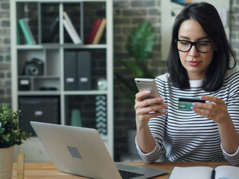 Junge Frau mit Brille macht eine online Überweisung mit der Kreditkarte und verwendet dafür Laptop und Handy