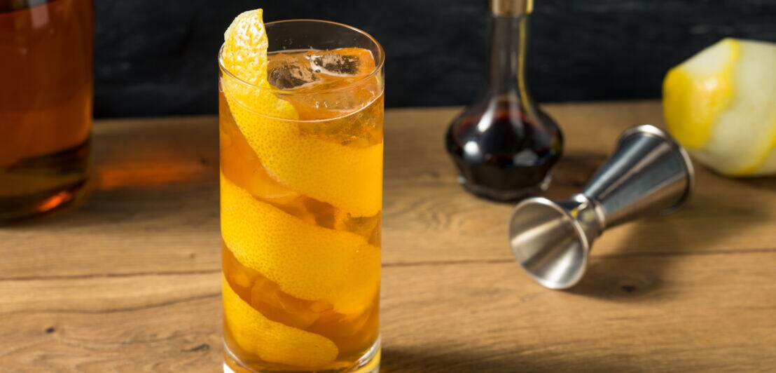 Cocktail mit Zitronenschale auf Holztisch, daneben Zutaten und Messbecher