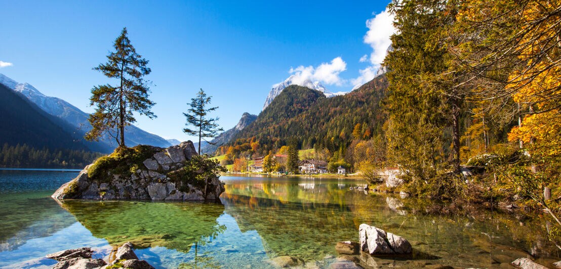 Ein See mit glasklarem Wasser mit Steinen, umgeben von herbstlichem Wald, im Hintergrund Bergpanorama