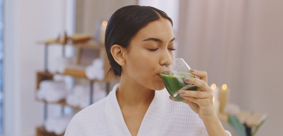 Frau in Bademantel, die mit geschlossenen Augen aus einem Glas mit grünem Inhalt trinkt
