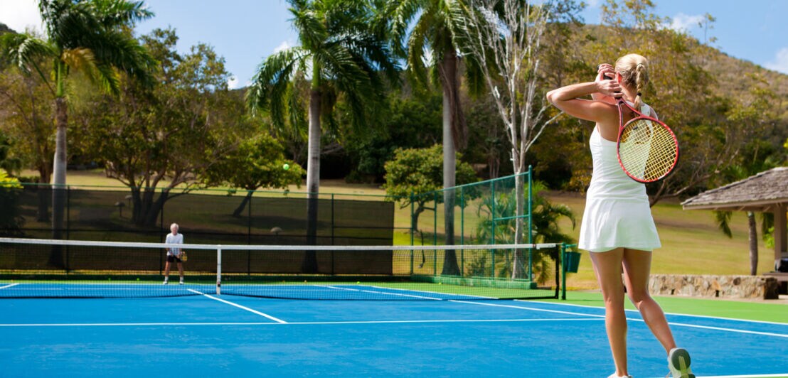 Zwei Personen spielen auf einem sonnigen Platz umgeben von Palmen Tennis.
