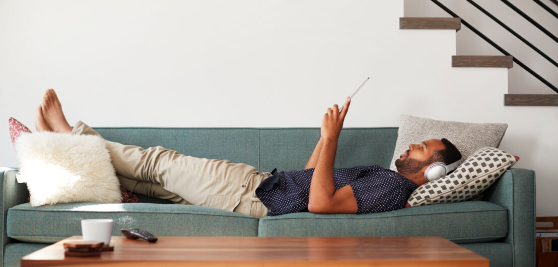 Ein Mann mit Kopfhörern, der auf ein Tablet schaut, liegend auf einem Sofa