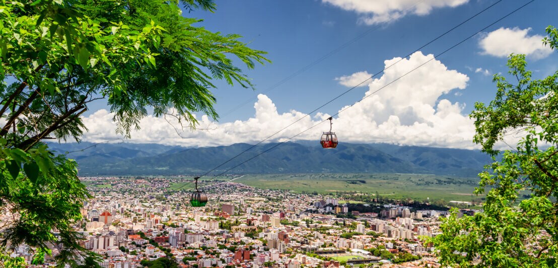 Panoramablick auf eine Seilbahn mit zwei Gondeln, die über der Stadt Salta einen Berg hinauffahren, im Hintergrund hügeliges Umland