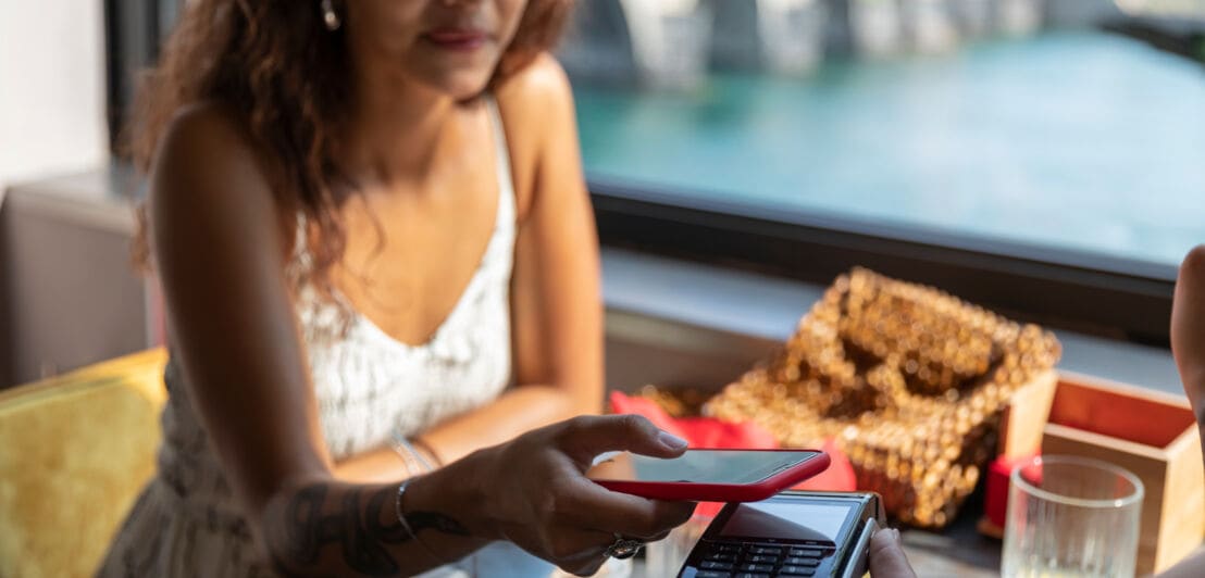 Eine junge Frau im Sommerkleid bezahlt kontaktlos mit ihrem Smartphone in einem Café an einem Fluss