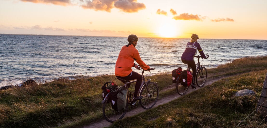 Rückansicht von zwei Personen auf Fahrrädern, die auf einem schmalen Radweg eine Küste bei Sonnenuntergang entlangfahren