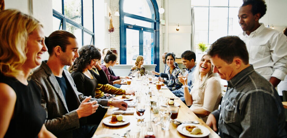 Eine Personengruppe sitzt beim Essen an einem langen Holztisch in einem kleinen Restaurant.