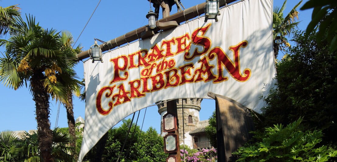Ein weißes Segel mit der Aufschrift Pirates of the Caribbean an einem großen Schiffsmast am Eingang einer Attraktion in einem Themenpark