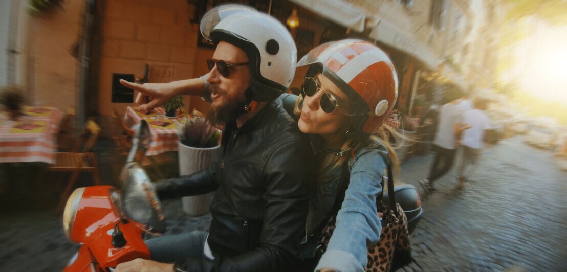 Ein junges, stylisches Paar mit Helmen fährt auf einem roten Motorroller durch eine schmale Straße mit Restaurants
