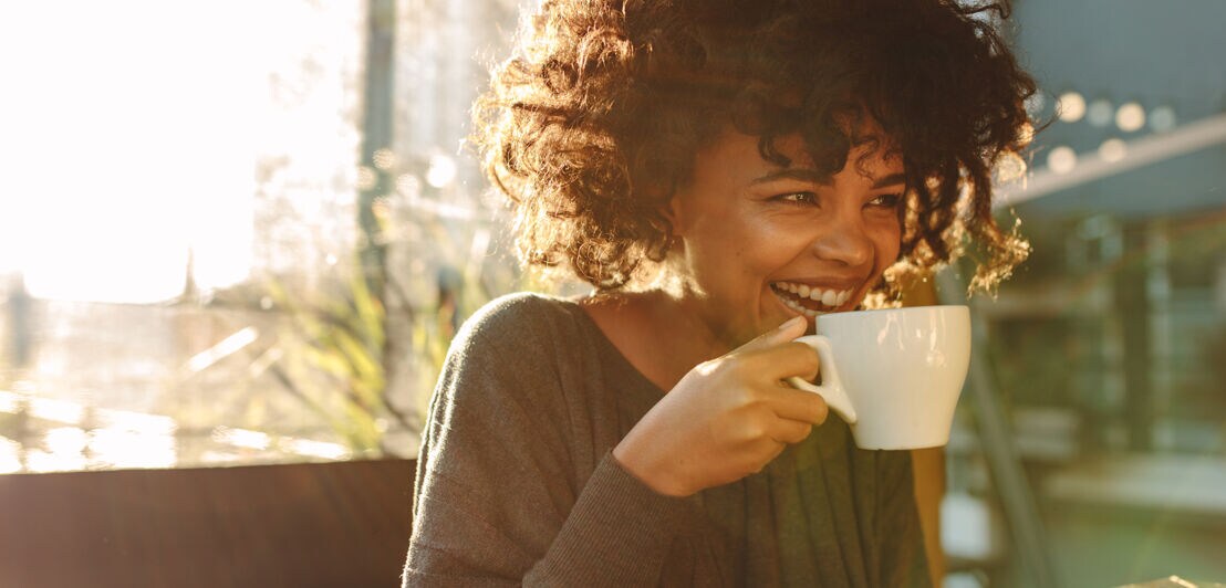 Smiling woman holding a coffee cup close to her face sitting inside a coffee shop. Curly haired woman drinking coffee.