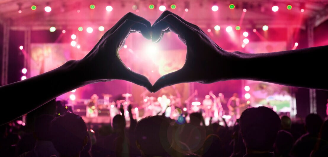 Zwei Hände formen ein Herz im Publikum vor einer Konzertbühne.