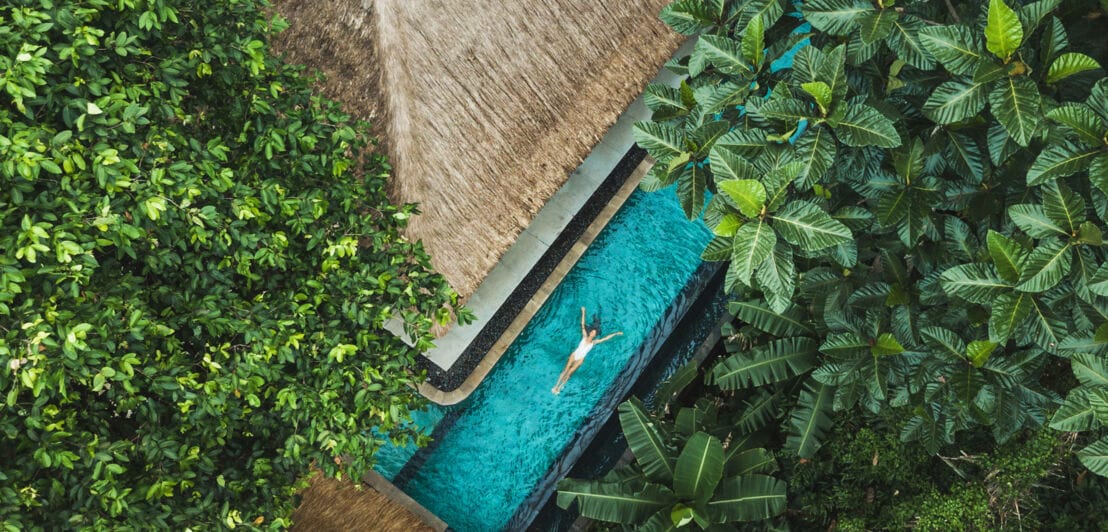Eine Frau schwimmt in einem Pool an einer Villa in tropischer Umgebung, aus Vogelperspektive