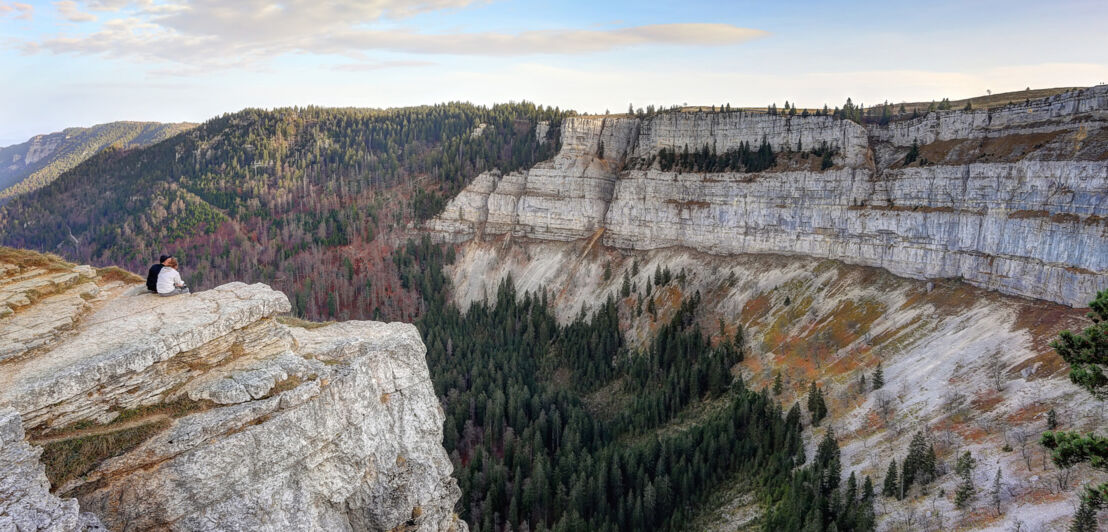 Zwei Personen sitzen auf einem Felsvorsprung vor einer gigantischen, senkrechten Felswand, umgeben von Waldlandschaft.