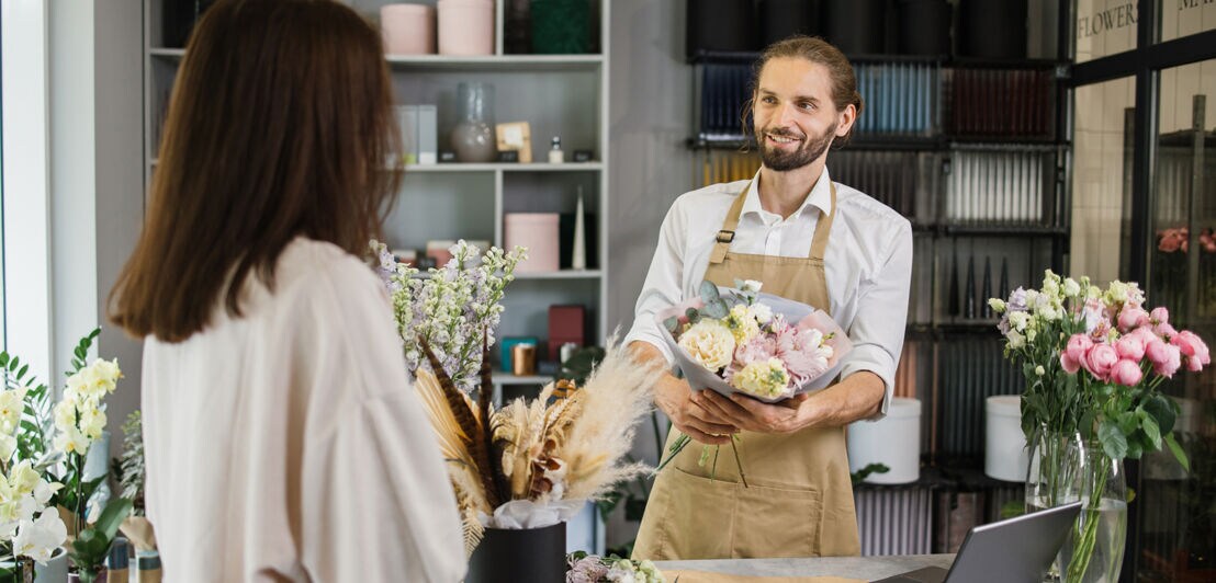 Attractive bearded man owner of flower shop advising female customer before purchase of flowers.