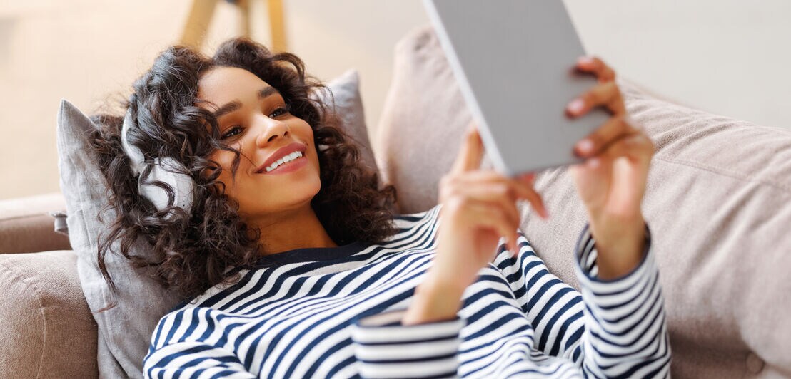 Delighted ethnic female smiling and browsing tablet while resting on couch and listening to music in headphones at home
