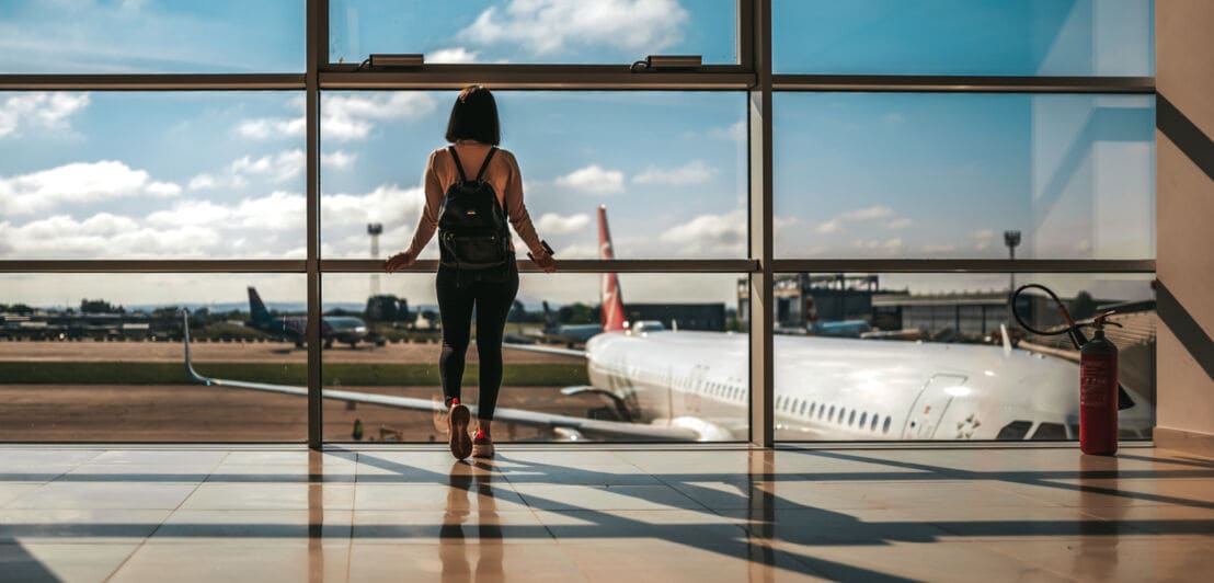 Eine Frau steht mit Rücken zur Kamera am bodenhohen Fenster am Gate eines Flughafens und blickt auf das Rollfeld.