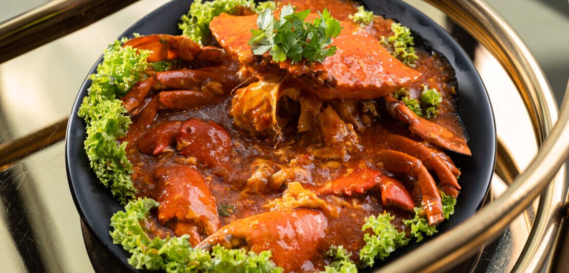 Eine gekochte Krabbe in roter Chilisoße auf einem schwarzen Teller, garniert mit Salatblättern.