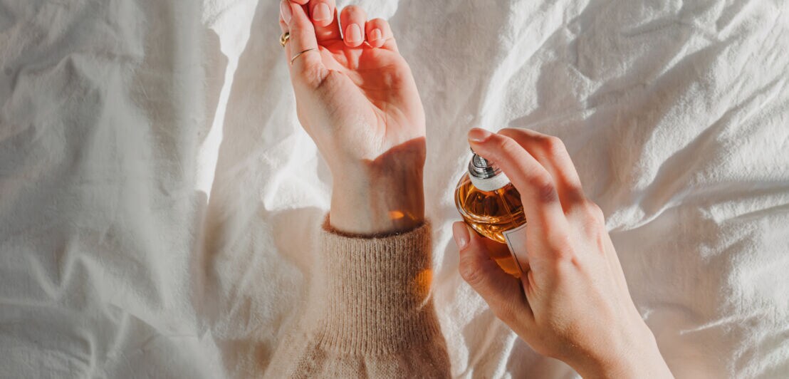 Nahaufnahme der Hände einer Person über einem weißen Bettlaken; in einer Hand hält sie eine orange-braune Parfumflasche, mit der sie das andere Handgelenk besprüht.