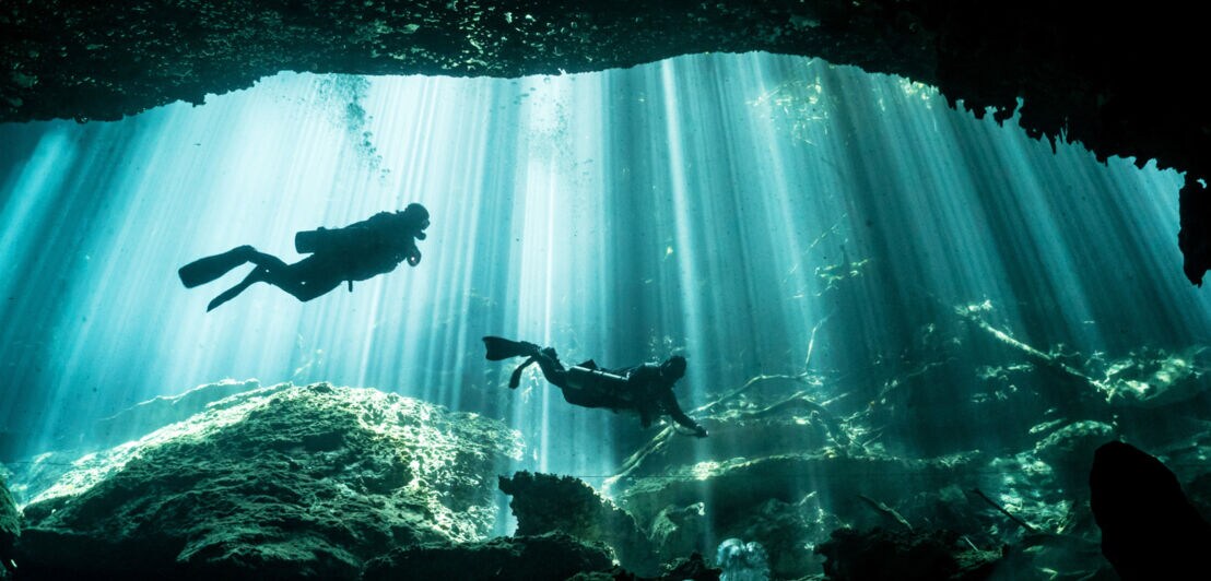 Zwei Taucher schwimmen durch Sonnenstrahlen in einer Unterwasserhöhle.