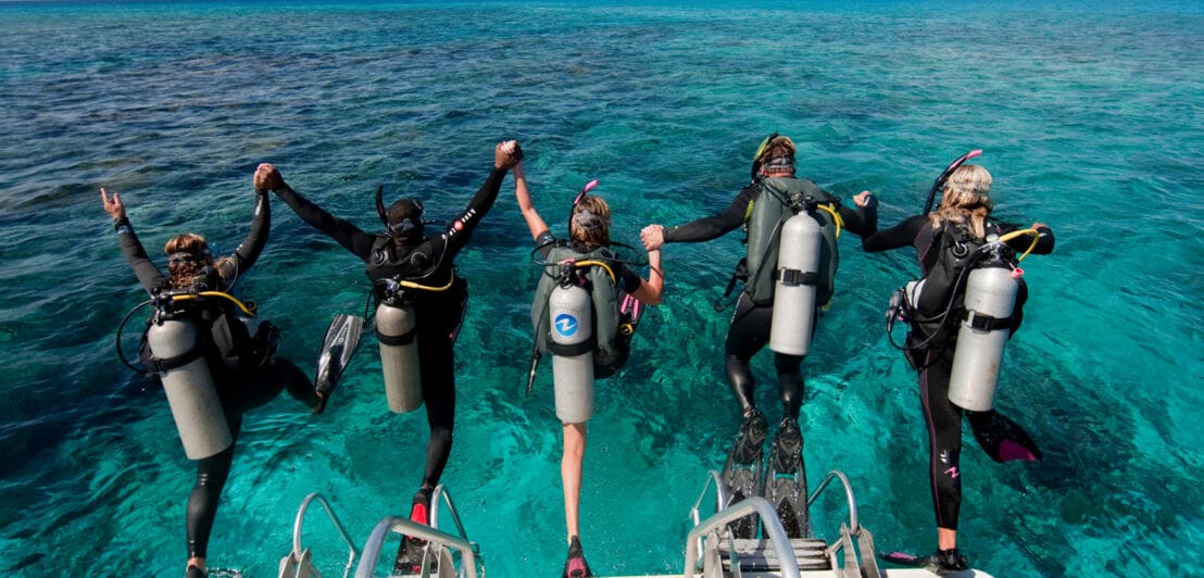 Fünf Taucher mit Sauerstoffflaschen auf den Rücken springen gemeinsam von einem Boot ins türkisblaue Meer.