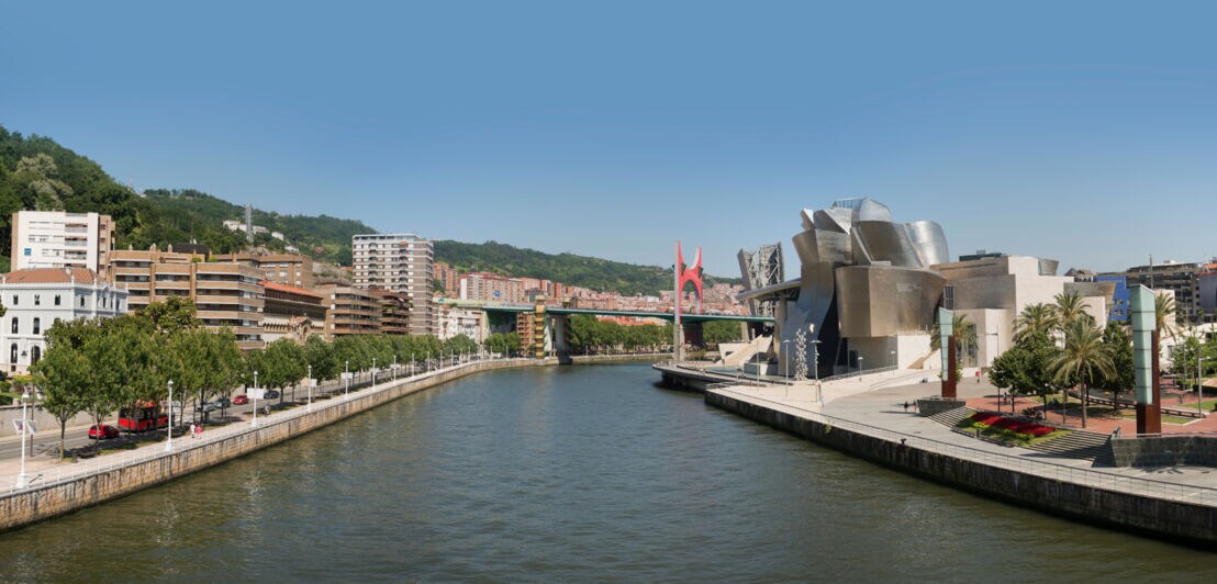 Das Guggenheim-Museum in Bilbao am rechten Ufer eines Flusses, weitere Häuser auf der anderen Flussseite.