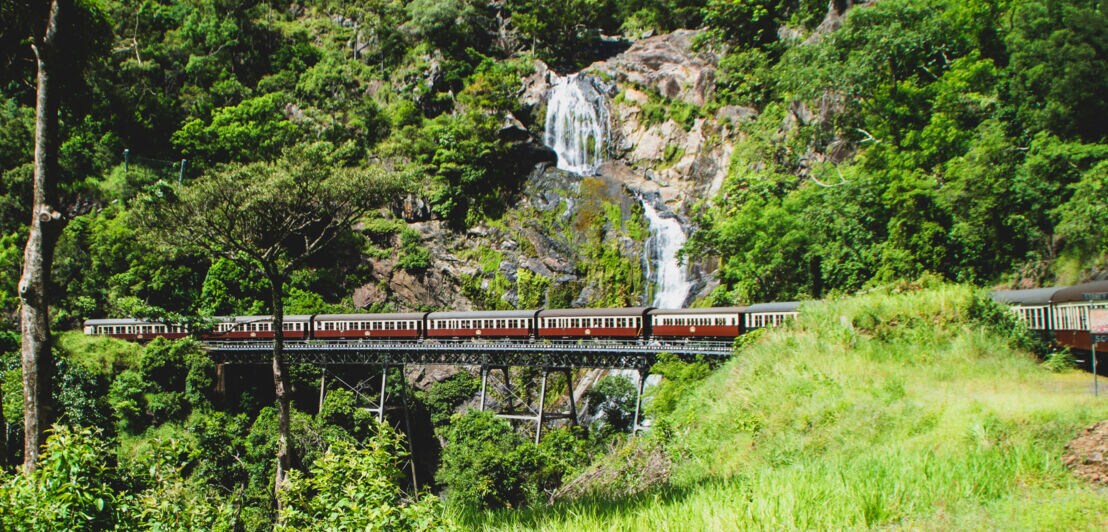 Eine historische Eisenbahn fährt über eine Brücke vor einem Wasserfall im Regenwald.
