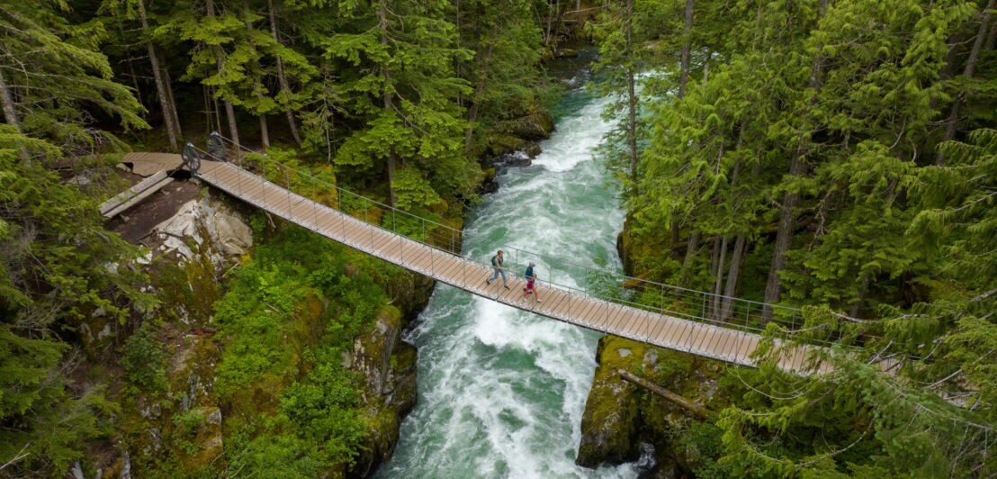 Luftaufnahme zweier Personen, die auf einer Hängebrücke einen wilden Fluss in einem Waldgebiet überqueren.