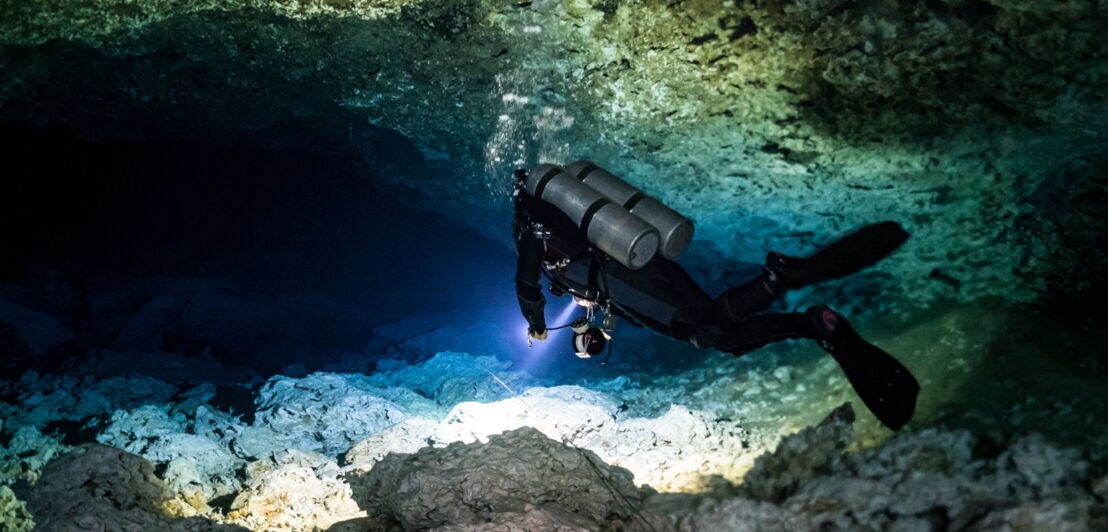Rückansicht einer Person in Taucherausrüstung, die den Grund einer Unterwasserhöhle mit einer Lampe anleuchtet.