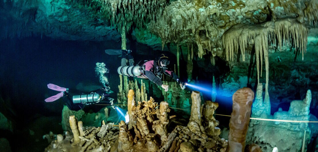 Zwei Personen in Tauchausrüstung, die durch eine Unterwasser-Tropfsteinhöhle mit Lampen schwimmen.