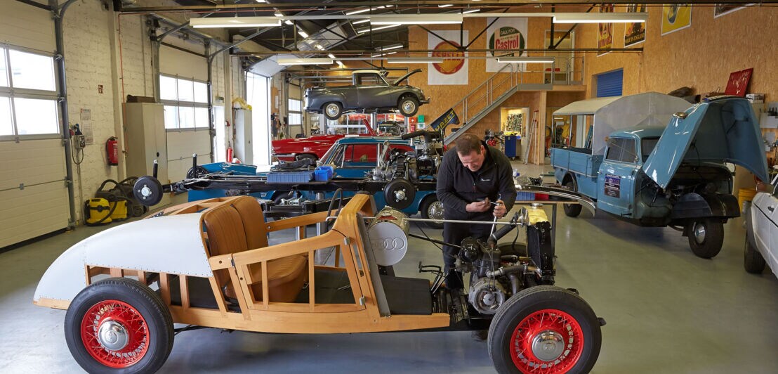 Ein Mechaniker arbeitet an einem Oldtimer mit Holzgestell in einer Werkstatt mit Ausstellungsraum.