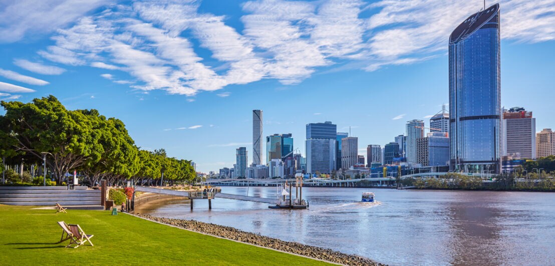 Grünfläche mit Liegestühlen am Brisbane River vor der Skyline der Stadt.