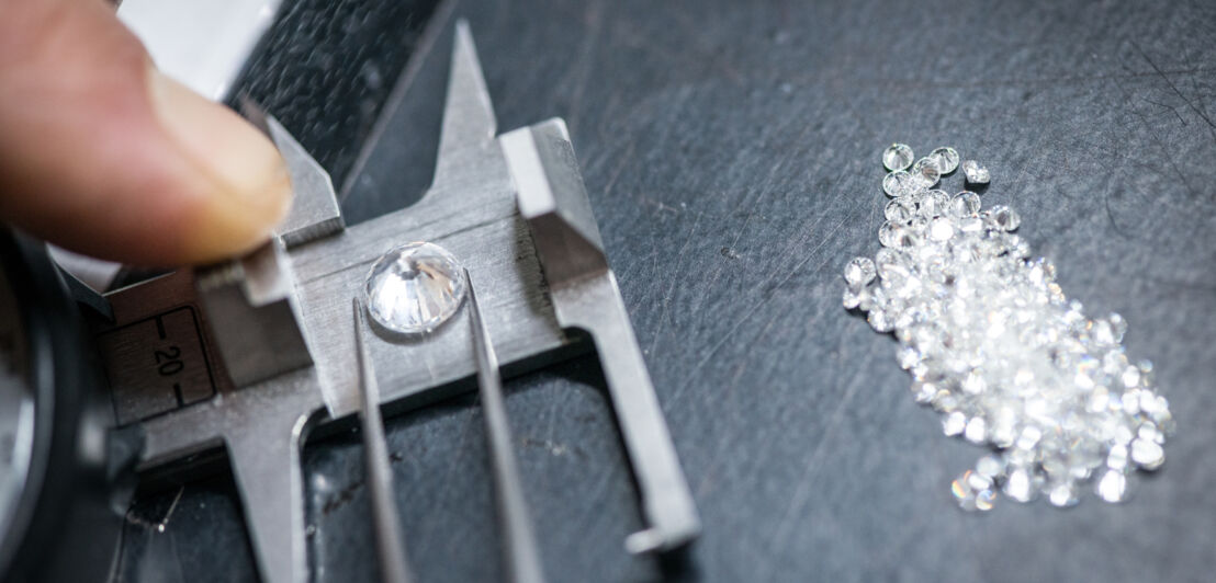 Ein Diamant liegt in einer Pinzette auf einer Waage, die von einem Finger gehalten