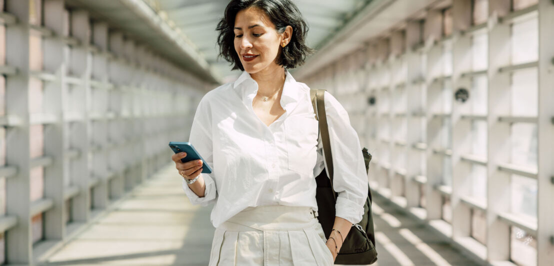 Eine elegant-lässige Frau in weißer Bluse und beiger Stoffhose schaut auf ihr Smartphone in einem hellen Durchgang.