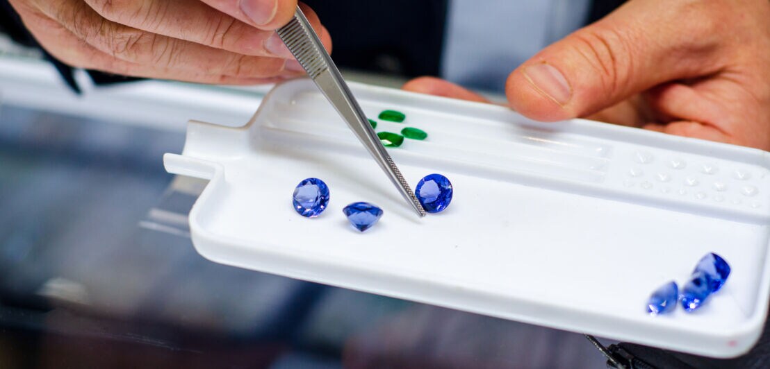 Mehrere blaue, geschliffene Edelsteine auf einem weißen Tablett, darüber eine Hand mit Pinzette.
