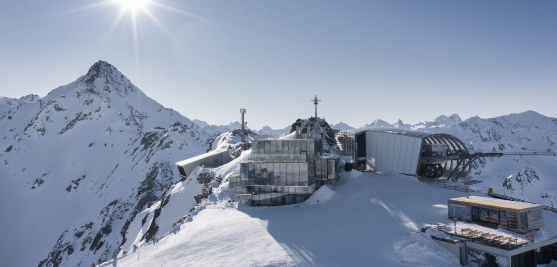 Kubistischer Gebäudekomplex mit verspiegelter Fassade neben einer Seilbahnstation auf einem schneebedeckten Gipfel bei Sonnenschein.