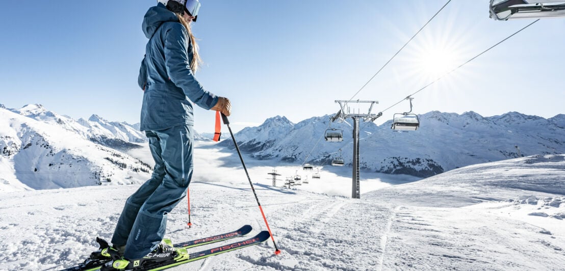 Rückansicht einer stehenden Skifahrerin auf einer Skipiste unter einem Sessellift vor Berglandschaft.