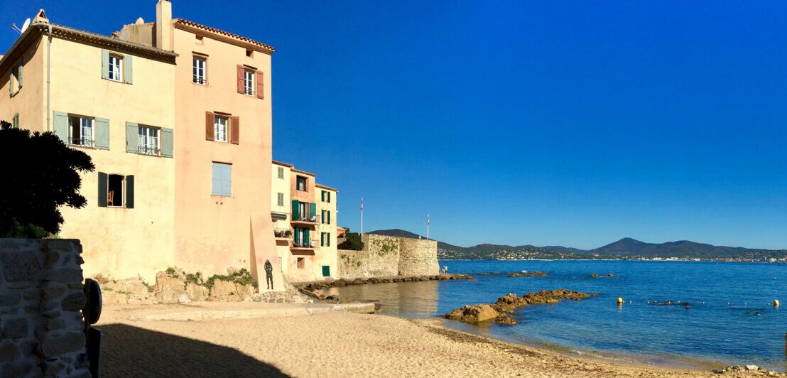 Kleine, menschenleere Strandbucht, umgeben von pastellfarbenen Häusern in mediterraner Umgebung.