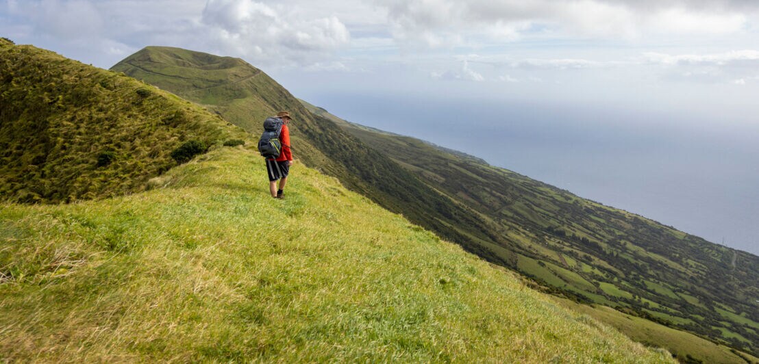 Eine Person mit Wanderausrüstung auf einem grün bewachsenen und steil abfallenden Vulkankegel der Azoreninsel São Jorge.