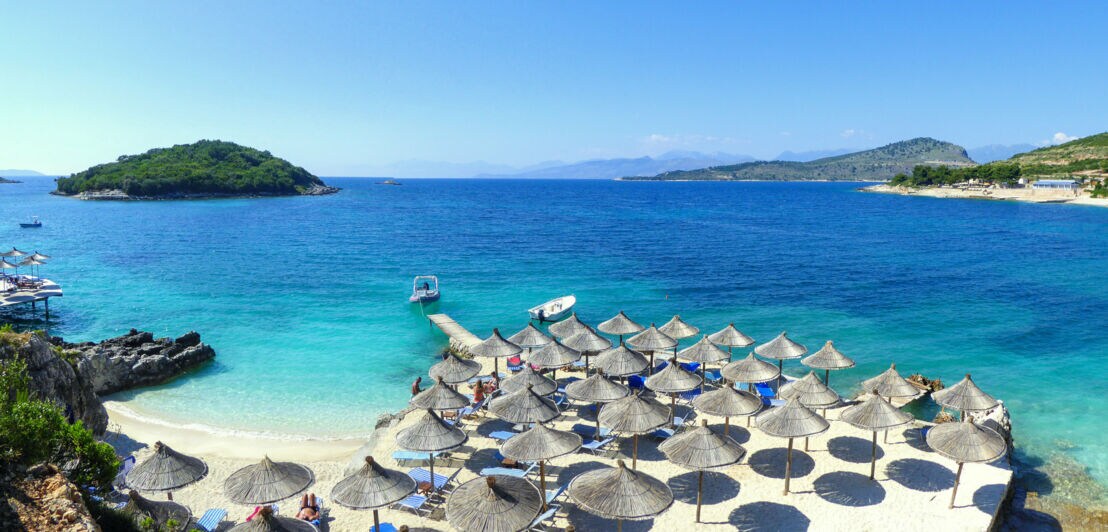 Strohschirme auf einem weißen Sandstrand in einer Bucht am türkislauem Meer.