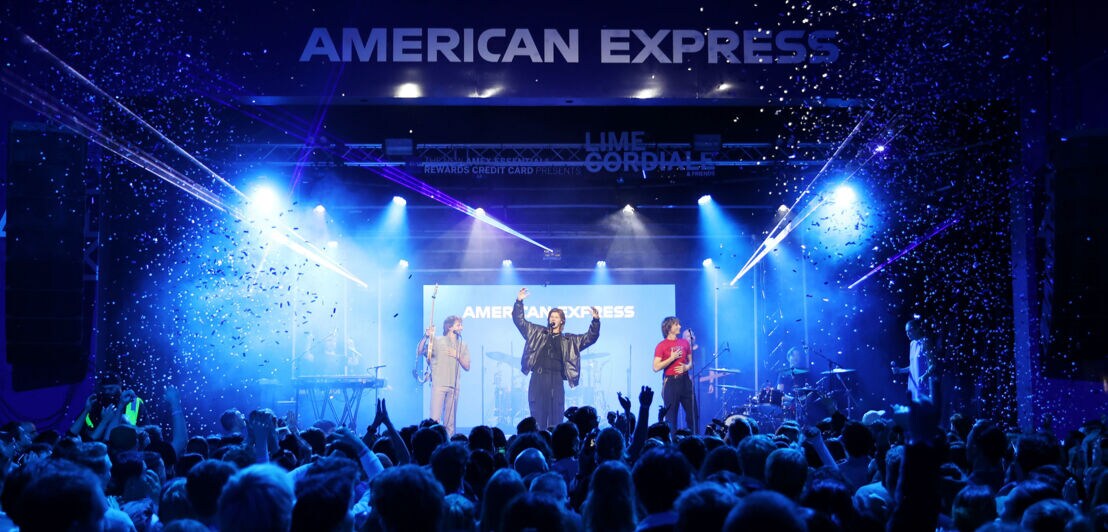 Eine Band auf einer blau beleuchteten Bühne mit American Express Schriftzug vor Publikum.