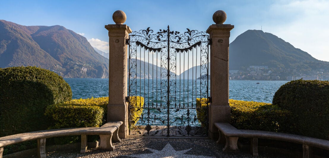 Ein schmiedeeisernes Tor in einer Parkanlage an einem See, im Hintergrund Berge.