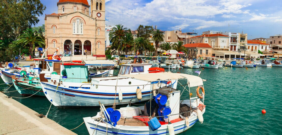 Mediterraner Ort mit Kirche an Hafenpromenade mit Fischerbooten im Vordergrund.