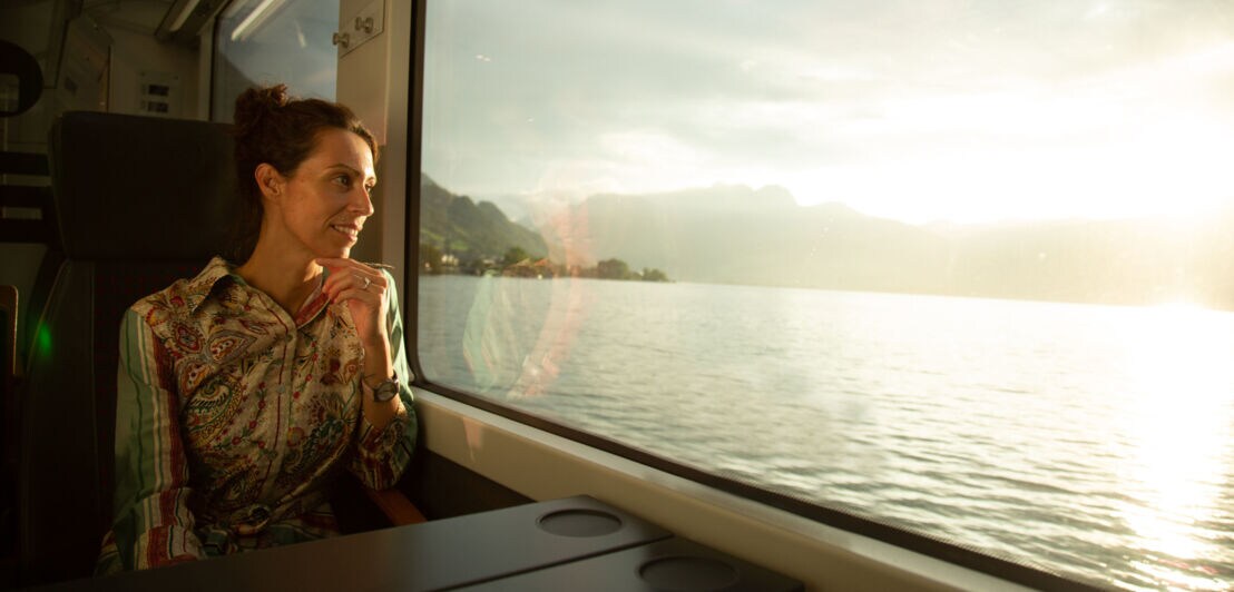 Eine Frau sitzt in einem Zug und blickt durch ein Panoramafenster auf einen See.