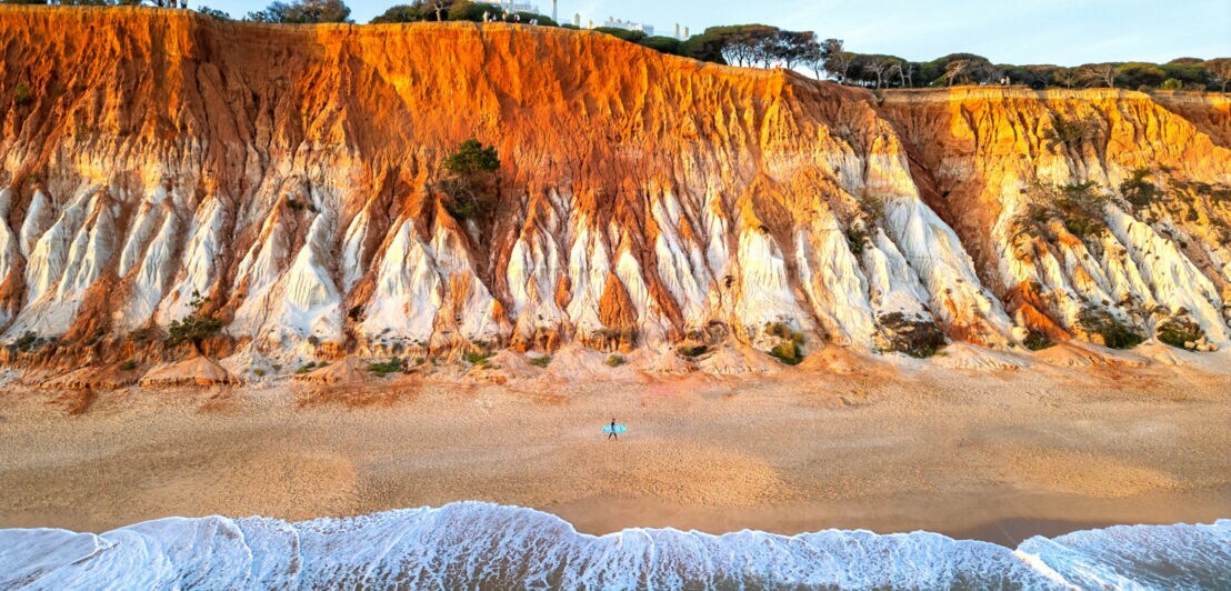 Hohe Klippen an der Praia da Falésia in Portugal, davor das Meer und der Strand, an dem eine Person mit Surfbrett entlangläuft.