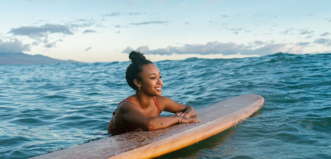 Eine lächelnde Frau hängt lässig mit den Armen an einem Surfbrett im Meer.