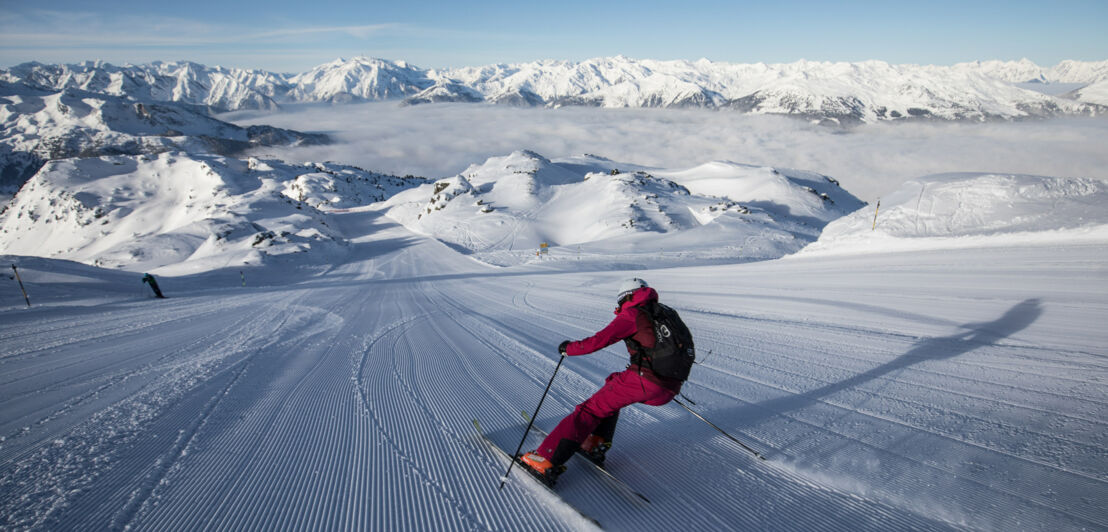 Rückansicht einer Skifahrerin auf einer breiten Piste vor schneebedeckten Gipfeln.