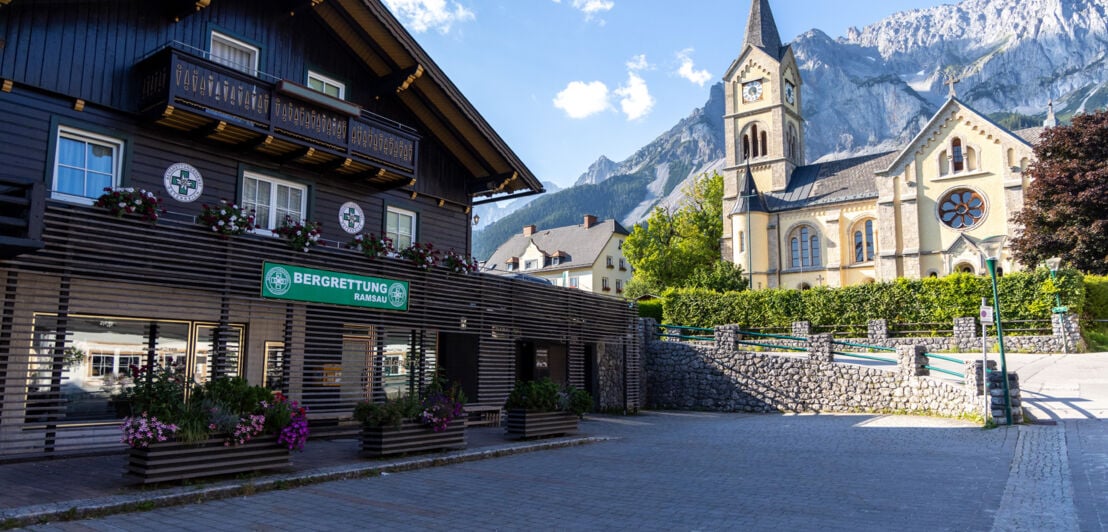 Bergrettungsstation in einem Holzhaus an einem Platz vor einer Kirche in einer Ortschaft mit Bergpanorama im Hintergrund.