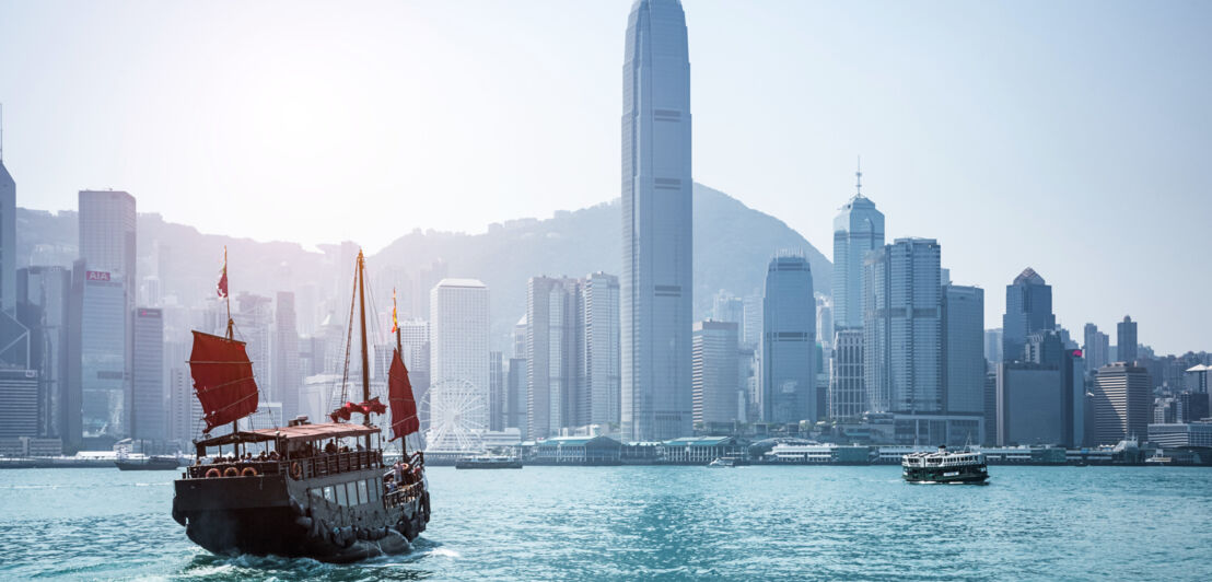 Chinesische Dschunke mit roten Segeln auf dem Wasser vor der Skyline Hongkongs.